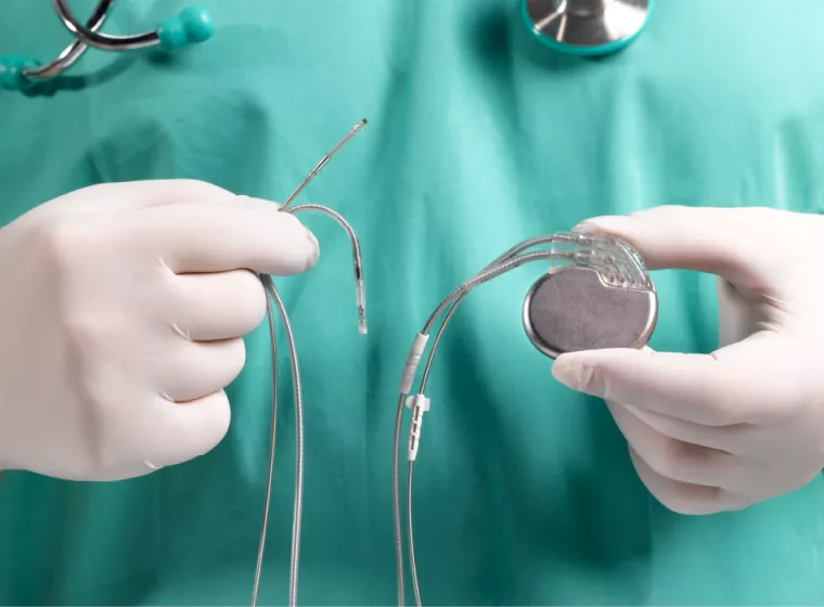 Gloved, medical professional in green scrubs with stethoscope around neck (shown from neck to mid torso) holding an implanted medical device